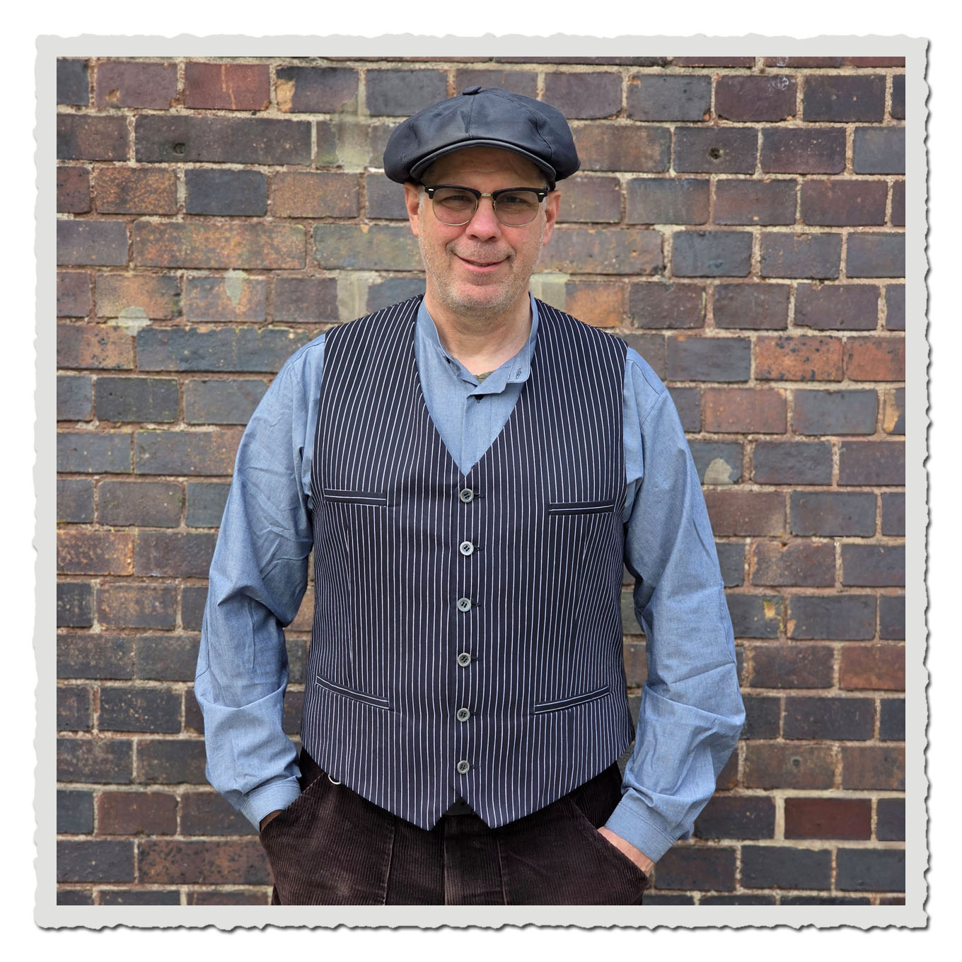 Mann in schwarzer gestreifter Weste und dunkler Mütze, Frontalansicht vor brauner Backsteinwand.